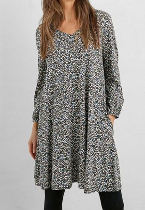 Kvinde iført langærmet, løs pasform, knælængde kjole med lille sort, hvid, blå og grøn abstrakt blomsterprint og sorte leggings.