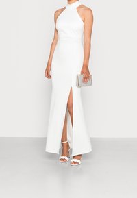 Robe blanche à encolure halter avec une fente latérale, au design ajusté, texture lisse. Associée à une pochette argentée et des sandales blanches à lanières.