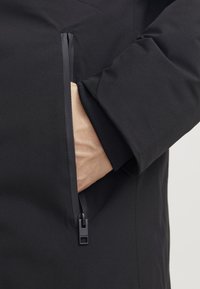 Sort isoleret jakke med et nærbillede af en lynlåslomme. Stoffet er glat, med en mat overflade og ensfarvet.