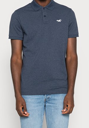 Mand iført en mørkeblå kortærmet polo med en hvid mågelogo på brystet og lyseblå jeans, stående mod en ensfarvet baggrund.