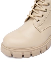 Botă din piele bej cu talpă groasă și texturată, ochiuri metalice și șireturi rotunde bej asortate, pe un fundal alb.