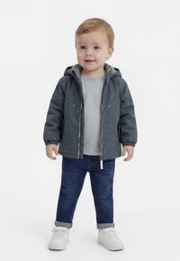 Kleinkind trägt eine dunkelgrüne, mit Reißverschluss versehene Kapuzenjacke, graues Hemd, blaue Jeans und weiße Turnschuhe und steht vor einem weißen Hintergrund.