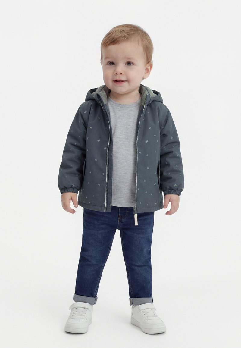 Kleinkind trägt eine dunkelgrüne, mit Reißverschluss versehene Kapuzenjacke, graues Hemd, blaue Jeans und weiße Turnschuhe und steht vor einem weißen Hintergrund.