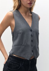 Grå skræddersyet vest med V-hals, der har seks knapper, let indsnævring i taljen og to forlommer. Stoffet har en glat, tekstureret overflade.