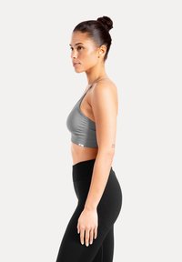 Brassière de sport grise avec fines bretelles, texture lisse et logo en bas, associée à un legging noir taille haute. Vue de côté.