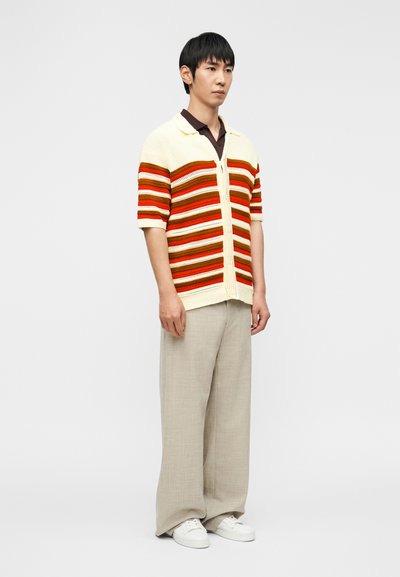 Άνδρας που στέκεται, φοράει μπεζ παντελόνι με φαρδιά πόδια, λευκά αθλητικά παπούτσια, καφέ πόλο και κρεμ κοντομάνικη ζακέτα με πορτοκαλί και καφέ ρίγες.