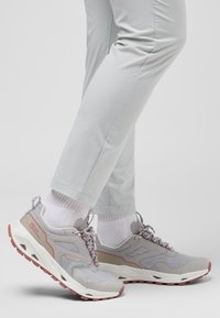 Chaussures de randonnée gris clair avec accents rouges, tige en mesh texturé, semelle en caoutchouc et détails réfléchissants. Associées à un pantalon clair et des chaussettes blanches.