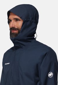 Navy waterproof jacket with a high collar and adjustable hood. Features a distinct logo on the sleeve and smooth fabric texture.