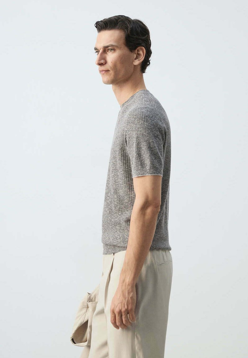Homme en pull gris à manches courtes et pantalon beige tenant une veste beige, debout de profil sur un fond clair uni.