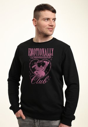 Muž s krátkými vlasy nosí černou mikinu s růžovou kreslenou postavičkou a nápisem „Emotionally Unavailable Club“, dívá se stranou.