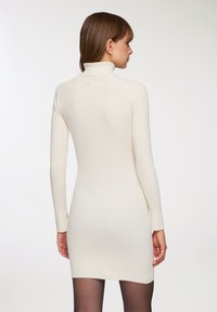 Vestito in maglia color crema chiaro con una texture a coste, dotato di un colletto alto e maniche lunghe, che termina sopra la parte superiore della coscia, con una silhouette aderente.