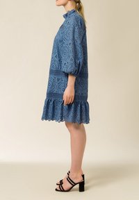 Vestido azul de ojo de perdiz con mangas abullonadas, dobladillo con volantes, detalles de encaje y un cuello alto, combinado con sandalias negras de tacón bloque con tiras.