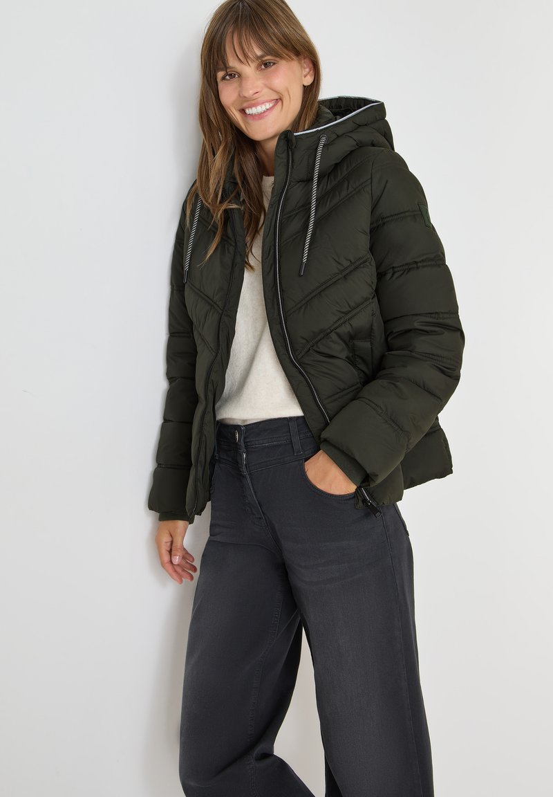 Mørkegrøn puffy jakke med hætte, med skrå quiltning, grå snoretræk og lynlås foran. Bæres over en lys sweater og mørke jeans.