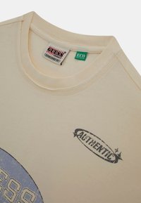 Kremowa bawełniana koszulka z okrągłym dekoltem. Posiada niebieską okrągłą grafikę oraz napis "AUTHENTIC" w kolorze czarnym. Widać metkę z kolekcji ekologicznej.
