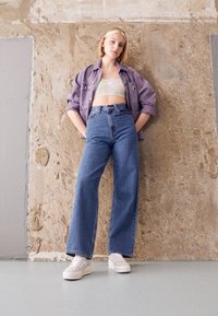 Světlofialová nadměrná bunda přes květovaný crop top, kombinovaná s vysokým pasem modrých džínů a bílými teniskami. Hladké textury, ležérní styl.