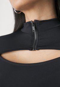 Top negro hecho de tela lisa, con un cuello alto y una cremallera frontal. Destacados cortes cerca de los hombros.
