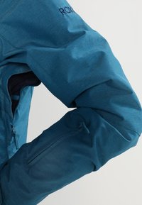 Blaue Stoffjacke mit strukturierter Oberfläche, ausgestattet mit einer Reißverschlusstasche am Ärmel und einer Logo-Stickerei in der Nähe der Schulter.