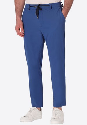 Uomo che indossa pantaloni blu slim-fit con coulisse nera e sneakers bianche, in piedi con una mano in tasca.