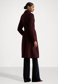 Cappotto in lana bordeaux con colletto, vita regolabile con cintura e maniche lunghe. Abbinato a pantaloni larghi scuri. Texture liscia, design minimalista.