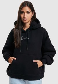 Sort hættetrøje lavet af blødt stof, med en frontlomme, snor i hætten og hvidt logo på brystet. Modellen bærer den med lyseblå jeans.