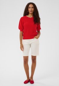 Pull en tricot rouge à manches courtes bouffantes, associé à un short blanc atteignant les genoux et des chaussures rouges tissées à enfiler.