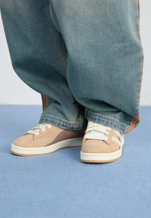 Baskets beiges en suède et tissu, avec des lacets blancs, des accents brun clair et une semelle texturée. Portées avec un pantalon en denim bleu ample.
