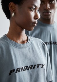 Camiseta de algodón gris con un destacado estampado negro de "PRIORITY". Cuello redondo, mangas cortas y textura suave. Dos modelos visibles, uno en primer plano.
