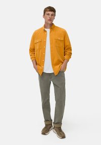 Langärmliges orangenes Cordhemd mit zwei Brusttaschen, kombiniert mit hellgrauen Hosen und braunen Sportschuhen, vor einem weißen Hintergrund stehend.