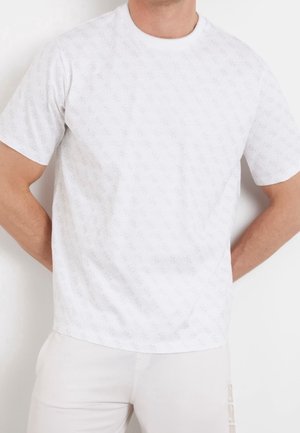 Homme portant un t-shirt blanc uni à manches courtes et un pantalon de couleur claire, debout les mains derrière le dos sur un fond blanc.