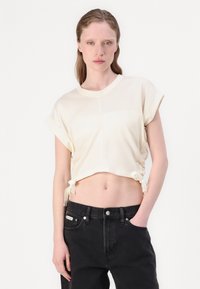 T-shirt cropped crema con maniche arrotolate, con lacci laterali. Indossato con jeans in denim neri a vita alta, creando una silhouette rilassata.