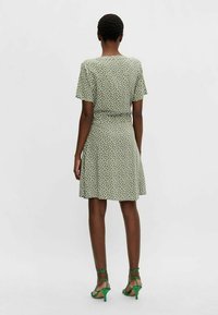 Robe verte à pois blancs, manches courtes, taille cintrée, jupe évasée et tissu lisse. Associée à des sandales vertes à lanières.