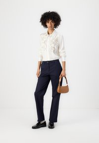 Weißes Hemd mit strukturierten floralen Verzierungen, kombiniert mit navyblauen Hosen. Schwarze Schuhe und eine kleine braune Handtasche runden den Look ab.