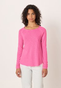 Ženska z kodrastimi lasmi, oblečena v svetlo roza majico z dolgimi rokavi in svetlo modre kavbojke, stoji ob enobarvnem svetlem ozadju.