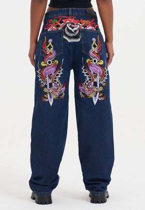 Jeans aus Denim mit bestickten lila Drachen- und Dolchmotiven auf den Gesäßtaschen, roter "Ed Hardy"-Schriftzug darüber, kombiniert mit schwarzem Oberteil und klobigen Stiefeln.
