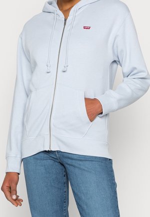 Person iført lyseblå hættetrøje med lynlås og forlommer samt rødt Levi's-logo, kombineret med blå jeans, hånd i lomme på en neutral baggrund.