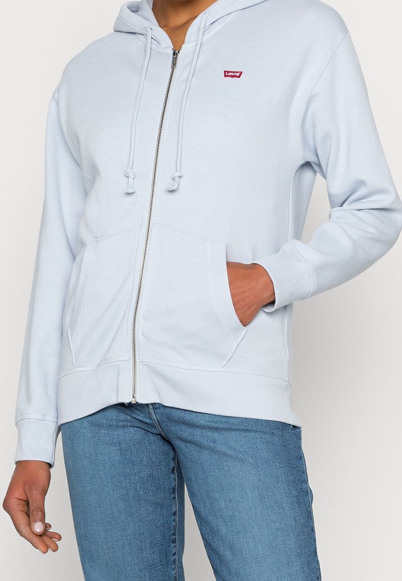 Personne portant un sweat à capuche zippé bleu clair avec poches avant et logo Levi’s rouge, associé à un jean bleu, main dans la poche sur un fond neutre.