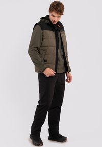 Wattierte Weste in Olivgrün mit schwarzem Kapuzen, Reißverschluss und Seitentaschen, über einem dunkelgrünen Pullover und schwarzen Hosen getragen.