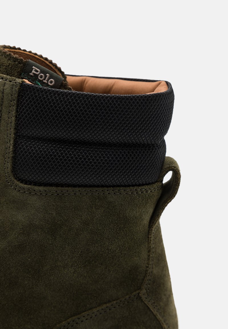 Grüne Wildlederstiefel mit einem strukturierten schwarzen Nylonkragen, ausgestattet mit einem braunen Innenfutter und einer kleinen Schlaufe an der Seite für ein einfaches Anziehen.