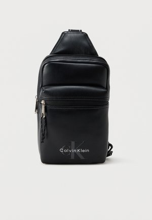 Czarna skórzana torebka typu sling Calvin Klein z przednią kieszenią na zamek i logo, mały, kompaktowy design, regulowany pasek na białym tle.