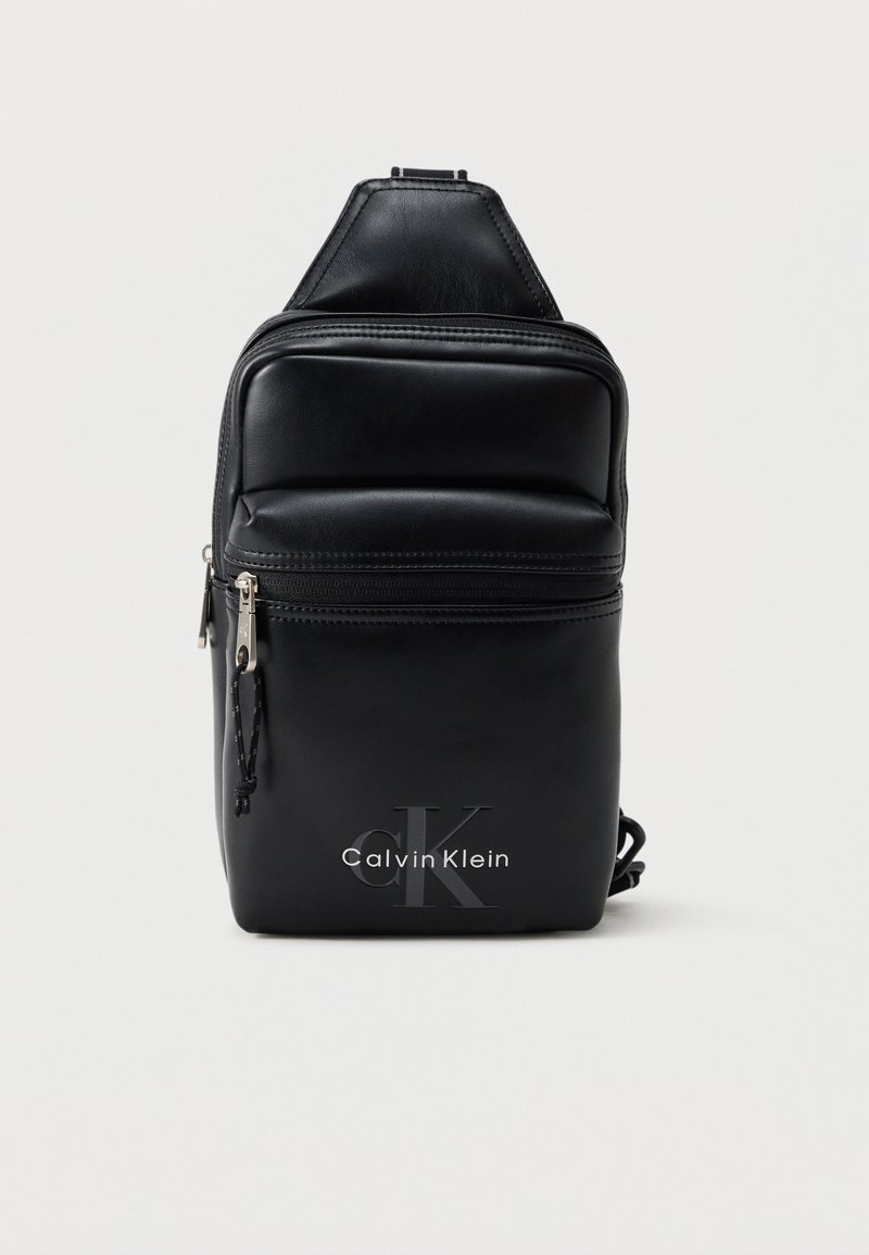 Μαύρο δερμάτινο τσαντάκι Calvin Klein με εμπρόσθια τσέπη με φερμουάρ και λογότυπο, μικρός συμπαγής σχεδιασμός, ρυθμιζόμενο λουρί σε λευκό φόντο.