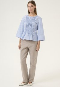 Blusa a righe blu e bianche con corpetto arricciato, maniche a tre quarti e orlo a peplo, abbinata a pantaloni beige a gamba dritta e scarpe basse.