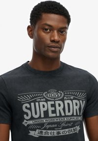 Zwarte katoenen T-shirt met een retro grijze grafische print met de tekst "SUPERDRY" en decoratieve accenten op de voorkant. Klassieke ronde halslijn.