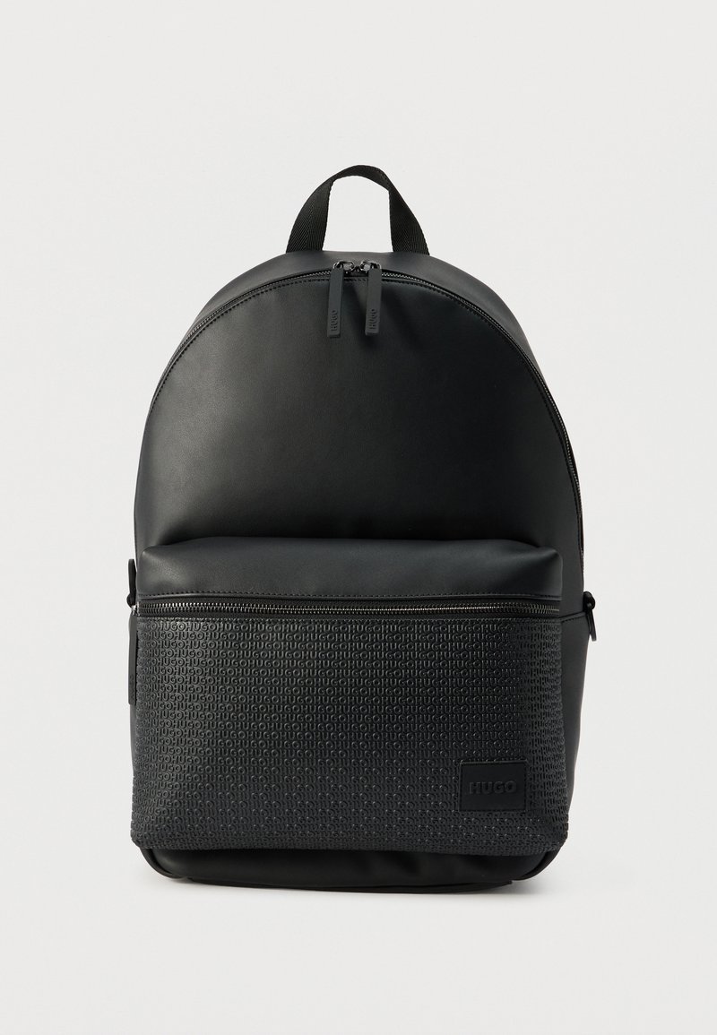 Sac à dos noir avec un dessus arrondi, une poche avant zippée embossée du texte "HUGO" répété, et deux curseurs de fermeture éclair sur le compartiment principal.