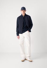 Navy bomber jacket over a navy sweater, white pants, and white sneakers with tan soles. The model wears a navy cap. Simple and clean design.
