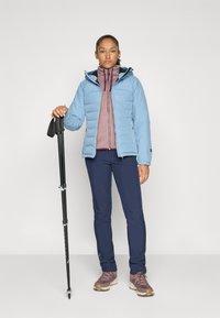 Kvinne i friluftsutstyr med lys blå jakke, lilla zip-up genser, marineblå bukser og tursko som holder en svart turstav.