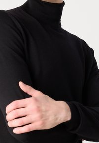 Schwarzer Rollkragenpullover mit geripptem Kragen, taillierter Passform, glatter Textur und langen Ärmeln, der die verschränkten Arme einer Person zeigt.