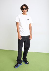 Hvid bomulds T-shirt med sort logo, parret med sorte denim jeans og sorte sneakers med blå såleaccepter. Grøn gulvbaggrund.