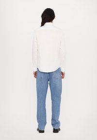 Personne aux longs cheveux foncés portant une chemise blanche à manches longues, un jean bleu et des chaussures noires, debout de dos sur un fond uni.