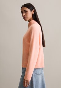 Jasnoróżowy sweter z dzianiny z prążkowanym wysokim kołnierzem i mankietami, w połączeniu z jasnoniebieską spódnicą dżinsową. Miękka struktura, luźny krój, prosty design.