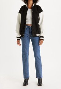 Chaqueta de varsity en blanco y negro con botones en el frente, puños acanalados y un diseño ajustado, combinada con jeans de pierna recta en azul claro y botas negras.